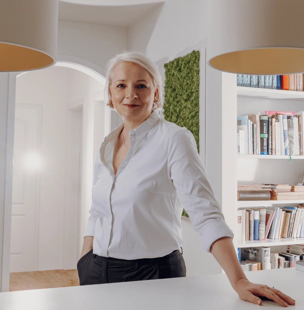 Eine Frau mit blonden Haaren steht in einem modernen Raum und lächelt. Sie trägt eine weiße Bluse und schwarze Hose und befindet sich vor einem Bücherregal.