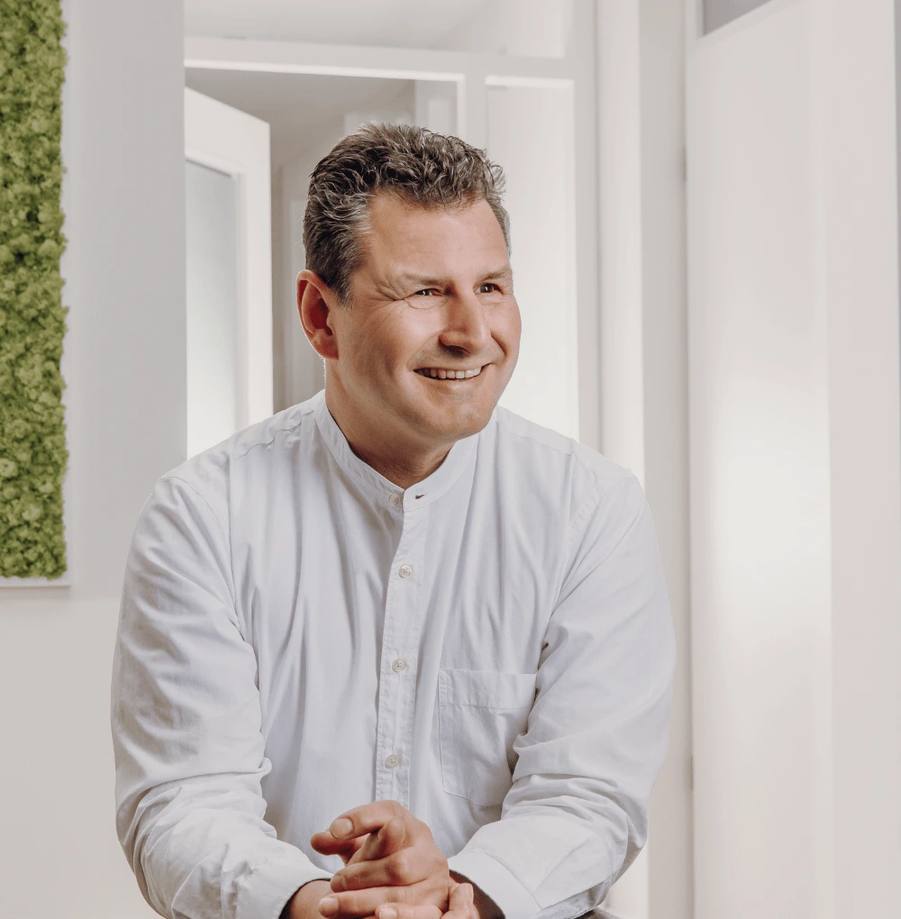 Lächelnde Person in einem hellen Raum mit weißen Wänden und grüner Pflanzenwand. Die Person trägt ein helles Hemd und sitzt entspannt.